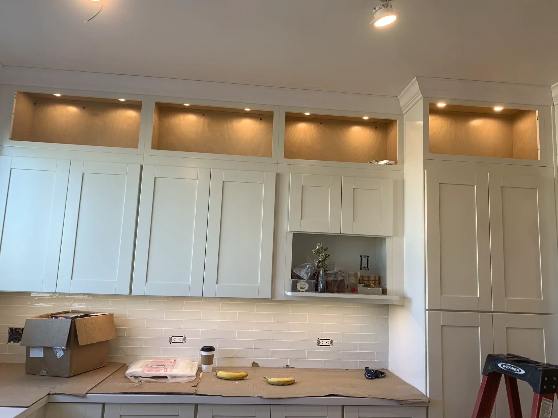 Partially completed kitchen with unpainted overhead cabinets, light fixtures, a tiled backsplash, and a red ladder to the right. Cardboard boxes and construction materials are scattered around.