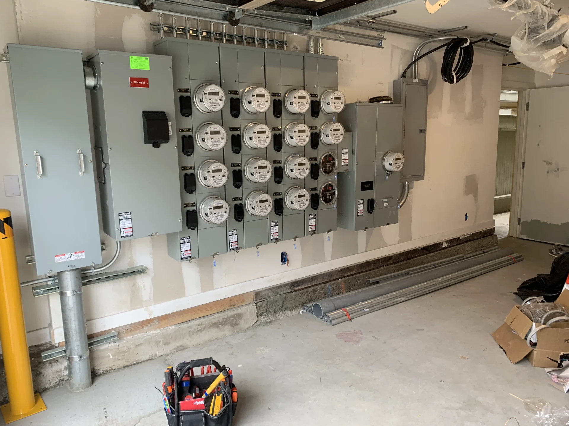 Service-Upgrades-1 - Aguilar Electric A room with multiple electric meters and electrical panels mounted on the wall. There are conduits and wiring materials on the floor, and a toolbox is placed nearby.