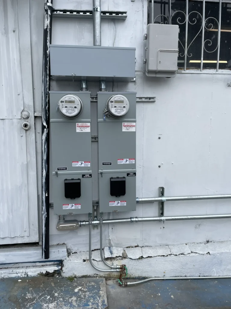 Service-Upgrades-4 - Aguilar Electric Two electrical meter boxes mounted on an exterior wall, with conduit pipes running below and to the side. Nearby is a door with peeling paint and a barred window.