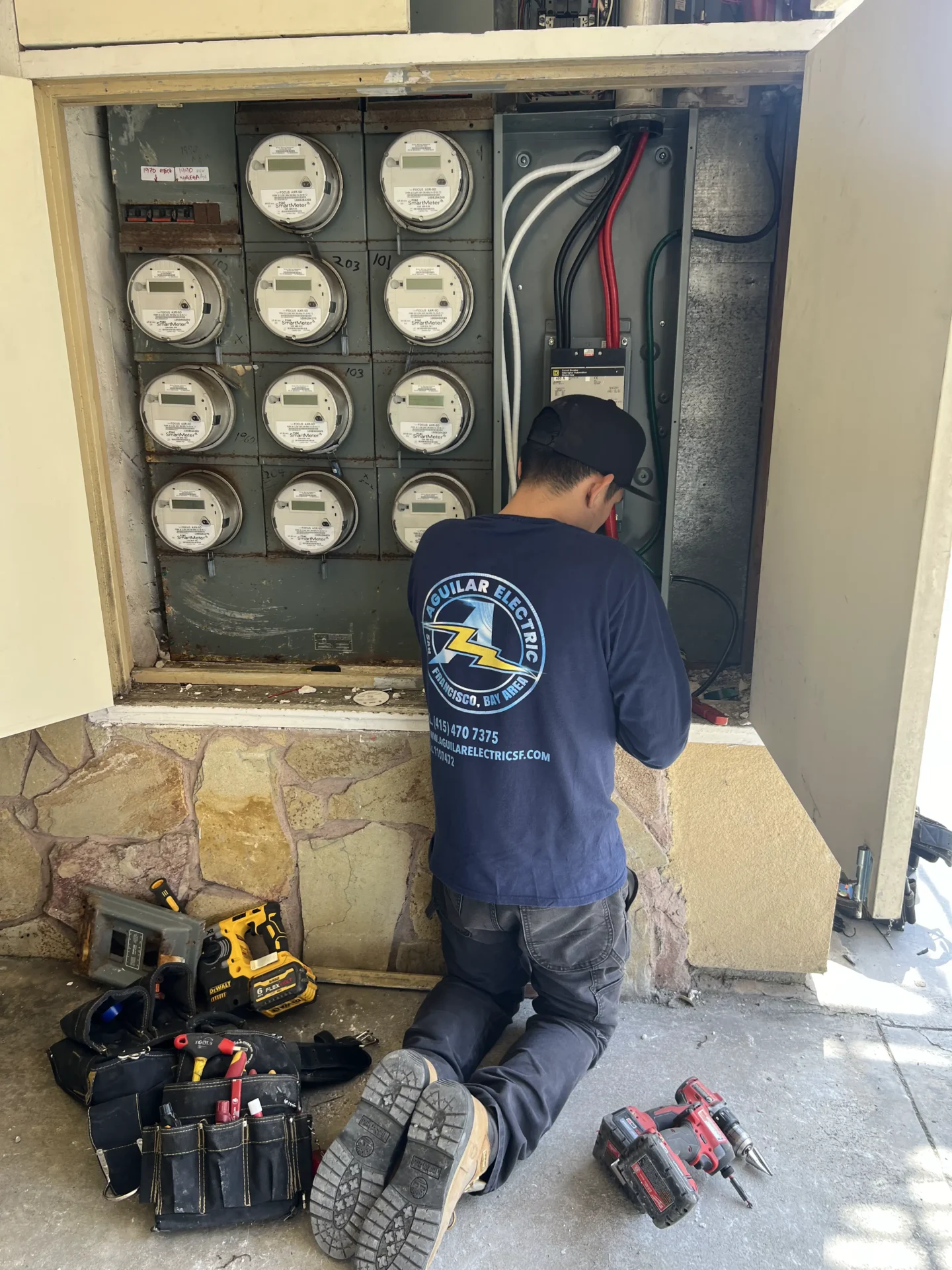 A worker wearing a "Zell Builders Electric" shirt kneels in front of an open electrical panel, performing maintenance. Tools and equipment are scattered around.