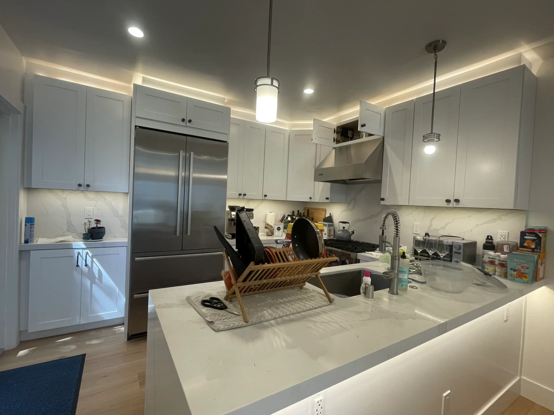 Modern kitchen with white cabinets, stainless steel appliances including a double-door refrigerator, and an island featuring a dish drying rack with dishes. Counters are cluttered with various items.